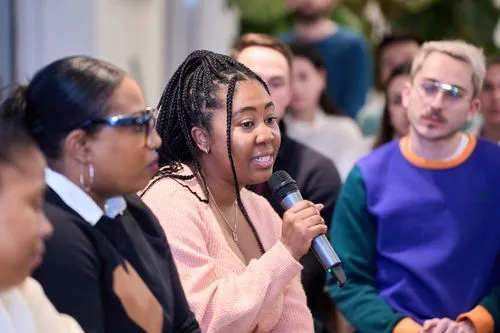 A woman with braided hair speaking into a microphone during a group discussion with attentive participants in the background.