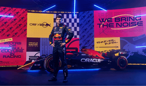 A Red Bull Racing driver poses confidently in front of a Formula 1 car and a vibrant backdrop celebrating 20 years of Red Bull culture in racing, featuring bold colors and slogans like "We Bring the Noise."