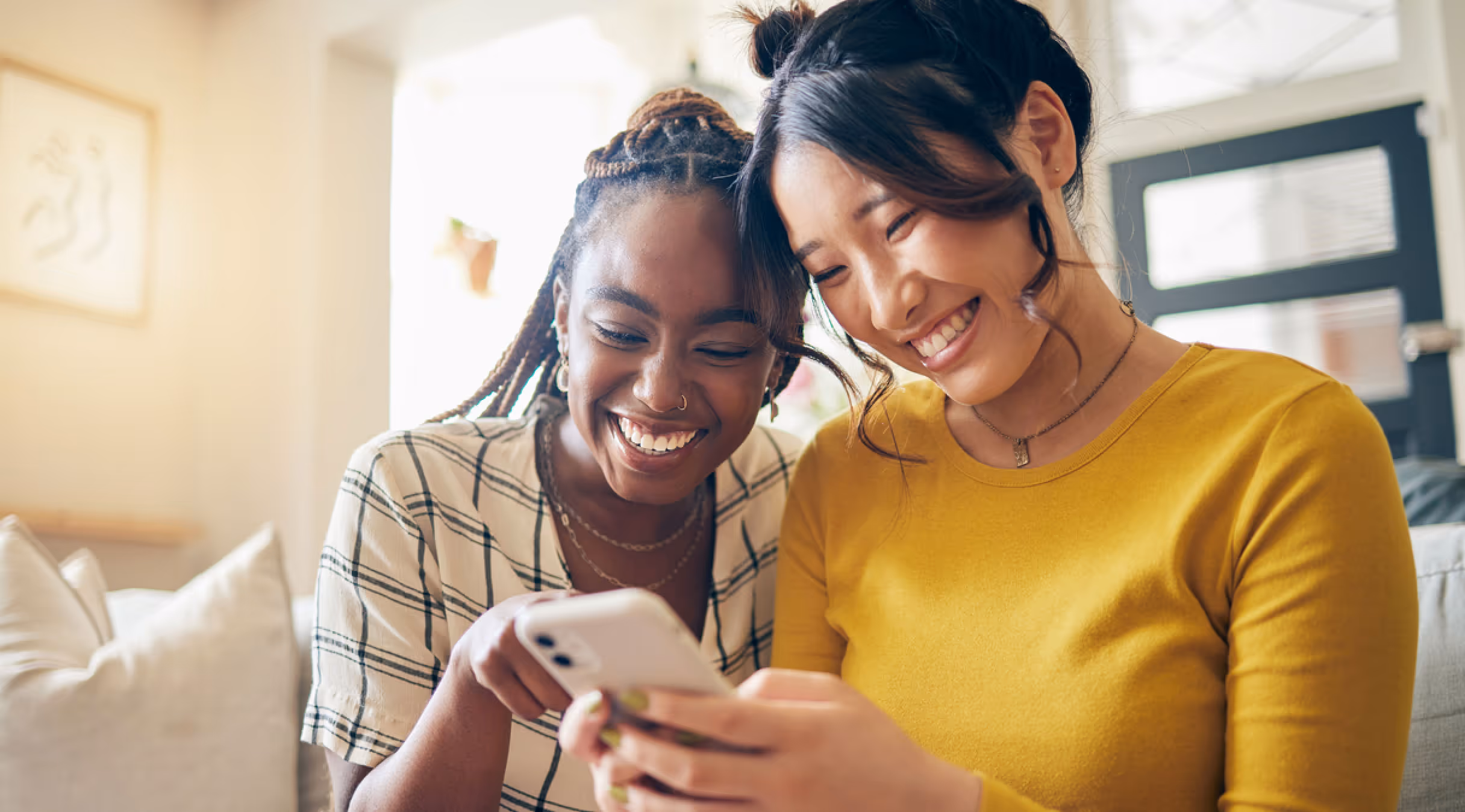 Two young women smiling joyfully while looking at a smartphone together indoors.