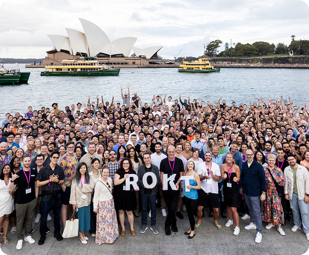 Large group of people posing together outdoors near water with Sydney Opera House and ferries in the background, some holding letters spelling ROKT.