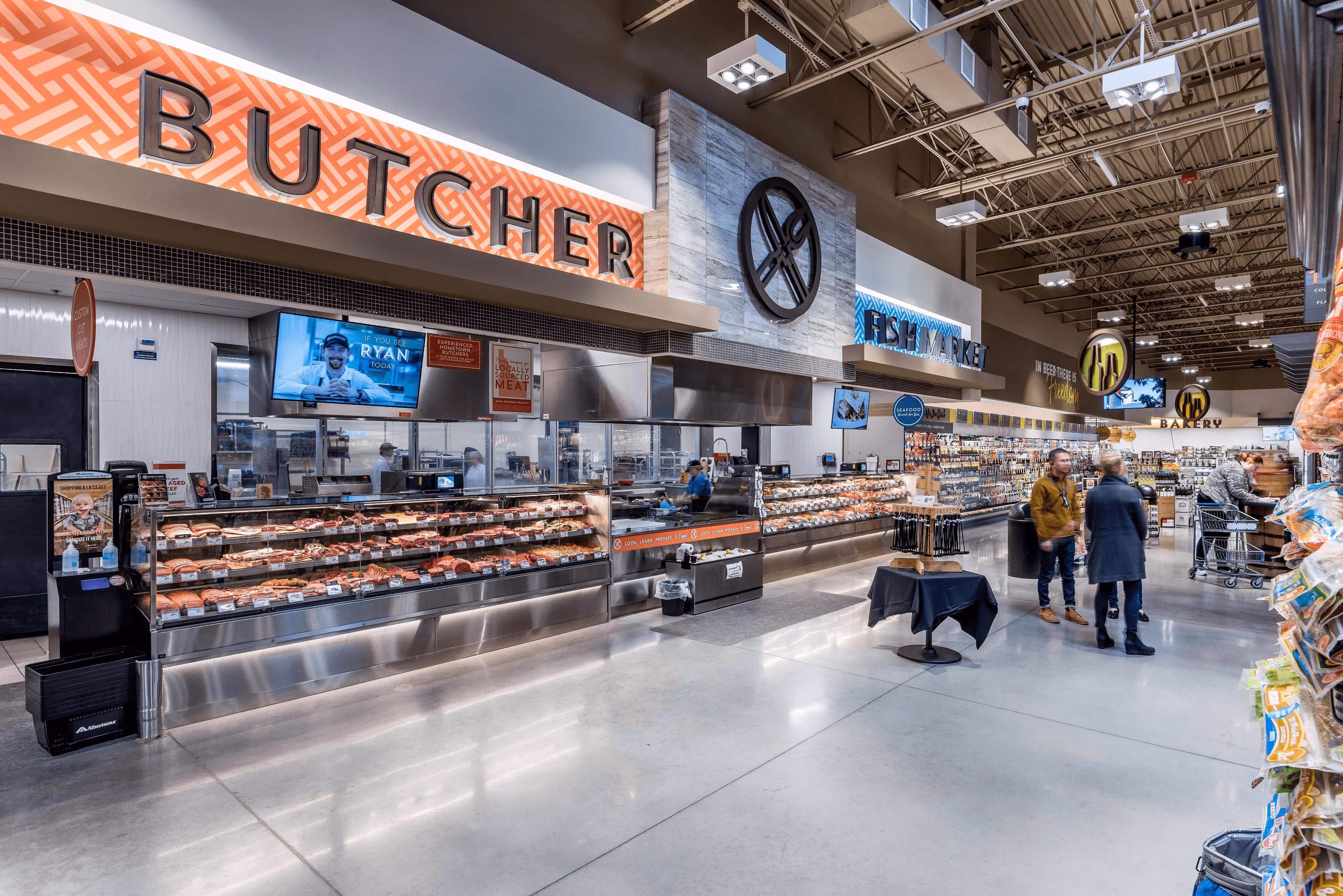 Interior of a modern grocery store featuring a butcher counter and fish market with customers shopping.