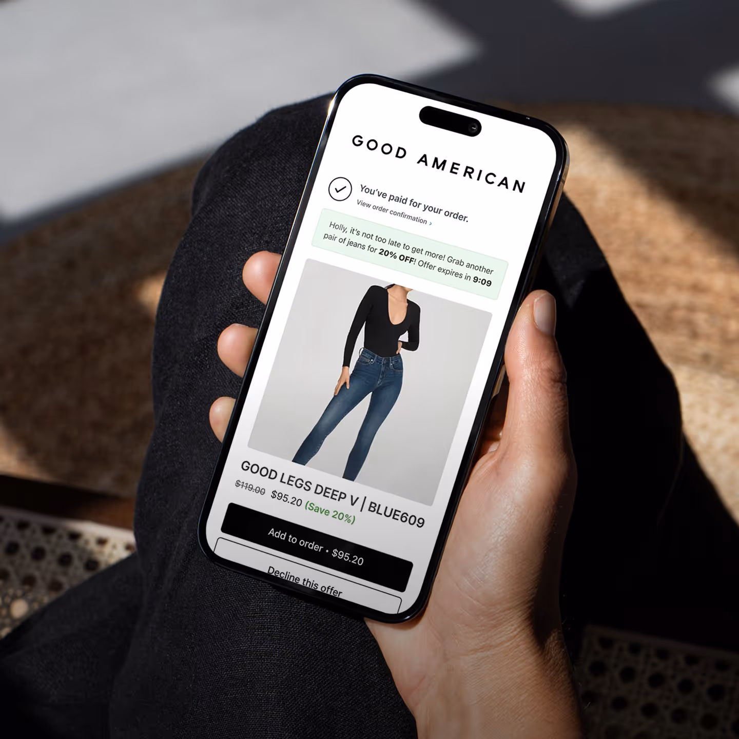 Hand holding smartphone showing Good American order confirmation with offer for 20% off a second pair of Good Legs Deep V jeans.
