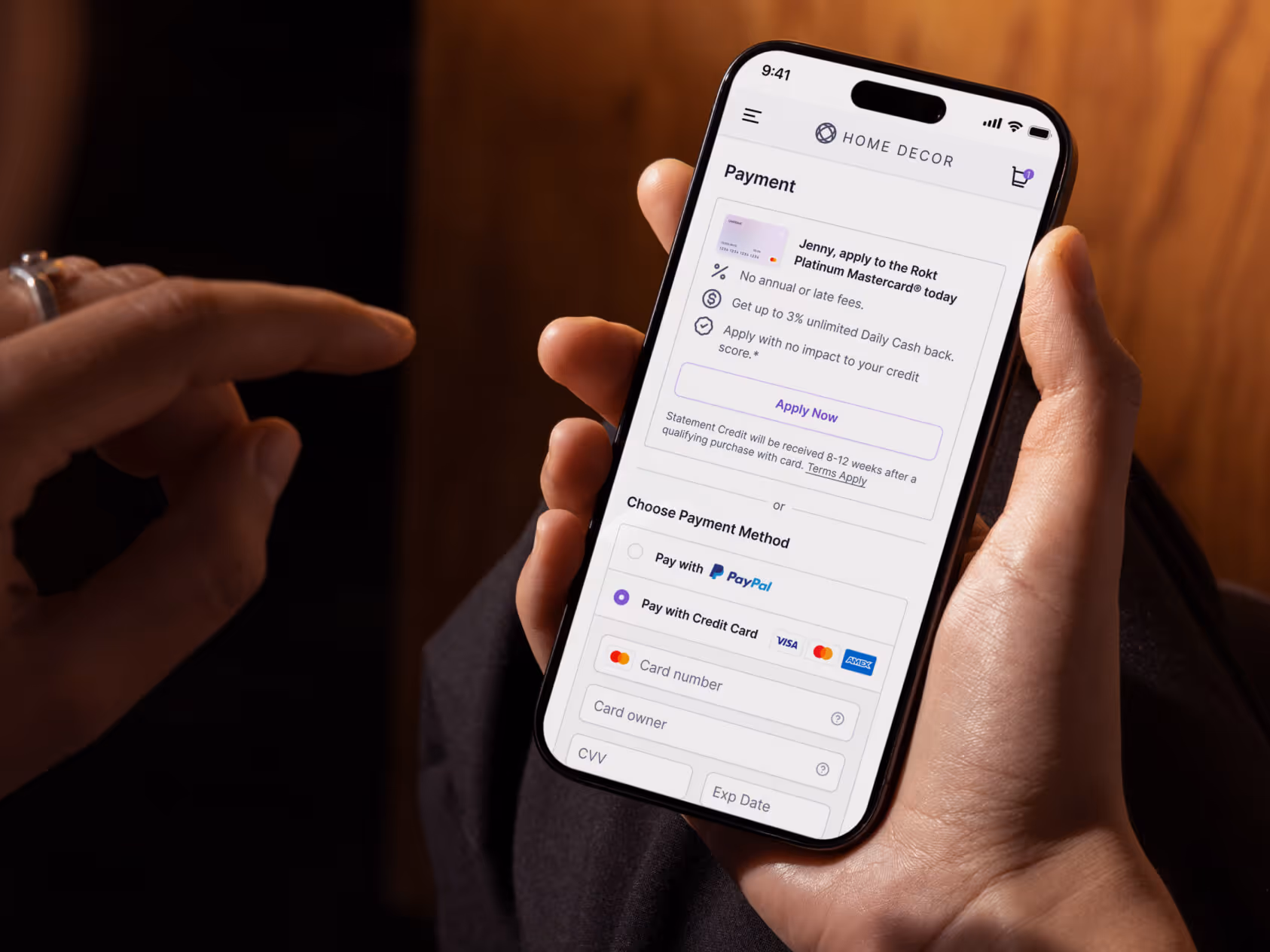 Une personne tient un smartphone alors qu'elle navigue sur la page de paiement d'une boutique en ligne appelée « Home Decor ». L'écran affiche une offre permettant de demander la carte Rokt Platinum Mastercard avec des avantages tels que l'absence de frais annuels et une remise en argent quotidienne allant jusqu'à 3 %. Ci-dessous, les utilisateurs peuvent choisir entre payer avec PayPal ou saisir les informations de leur carte de crédit. Le doigt du spectateur passe près de l'écran, ce qui suggère une interaction ou une décision imminente.