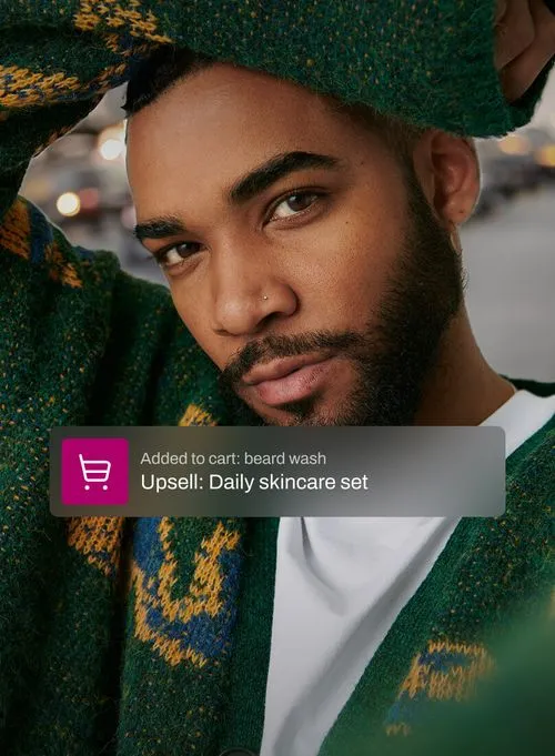 A man with a well-groomed beard and a cozy green sweater looks confidently into the camera. An overlay indicates he added beard wash to his cart, prompting an upsell for a daily skincare set, highlighting tailored product suggestions.