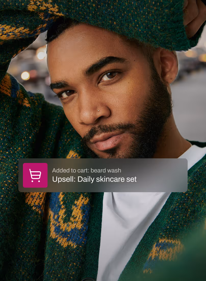 A man with a well-groomed beard and a cozy green sweater looks confidently into the camera. An overlay indicates he added beard wash to his cart, prompting an upsell for a daily skincare set, highlighting tailored product suggestions.