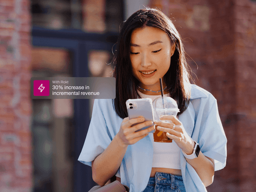 A young woman stands outside against a blurred brick background, smiling while looking at her smartphone. She holds a clear plastic cup with iced tea in one hand. Overlay text beside her reads “↑ 30% incremental revenue,” highlighting a mobile engagement benefit or business result.