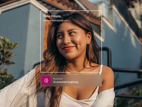 長い黒髪の若い女性は、明るい日差しの中で屋外で自信を持って微笑んでいます。オーバーレイには、「Rokt Brain — AI を活用したターゲティング」というラベルの付いたバナーとともに、彼女の名前とメールアドレス(「タリア・レイズ、taliareyes1992@gmail.com」)が表示されます。彼女はカラフルな建物に逆らって、フレンドリーでプロフェッショナルな雰囲気を醸し出しています。