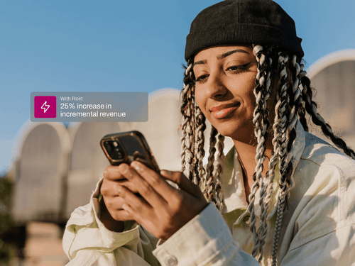 A woman with long twisted braids, wearing a black beanie and light-colored jacket, smiles slightly while looking at her phone outdoors under clear blue skies. A translucent overlay reads, “With Rokt: 30% increase in incremental revenue,” highlighting the business impact of the platform.