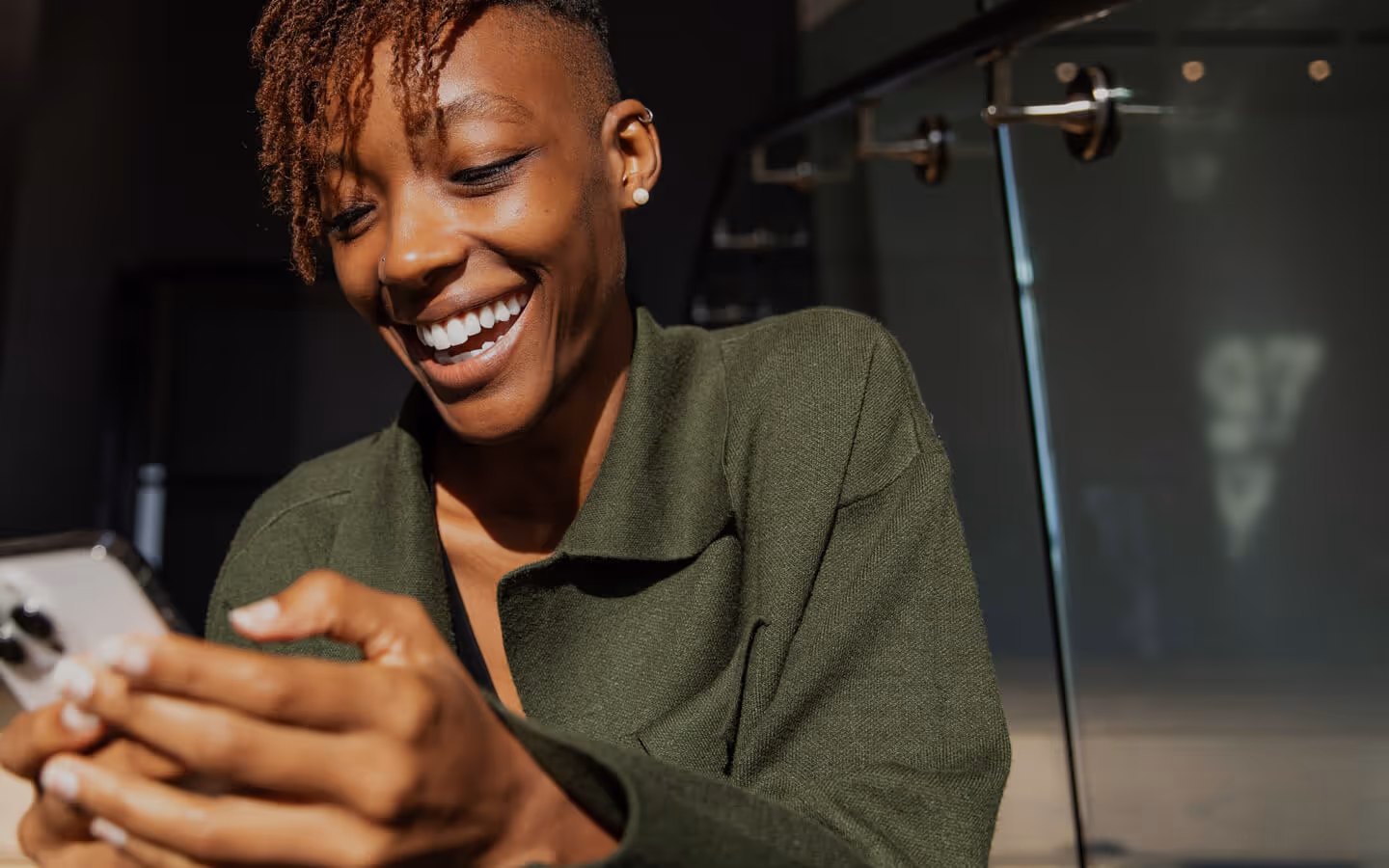 A woman with short natural hair and a green sweater laughs joyfully while using her smartphone indoors. The scene captures a moment of genuine connection or delight, enhanced by soft natural light and modern glass surroundings.