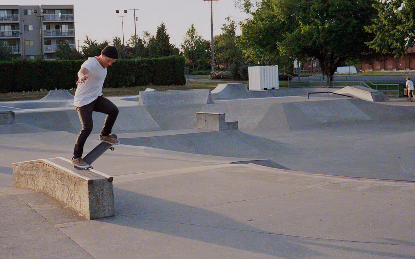 A skateboarder performs a trick on a concrete ledge at an outdoor skatepark surrounded by ramps, rails, and distant trees. The scene captures motion and balance during golden hour, with warm light casting shadows across the open space.