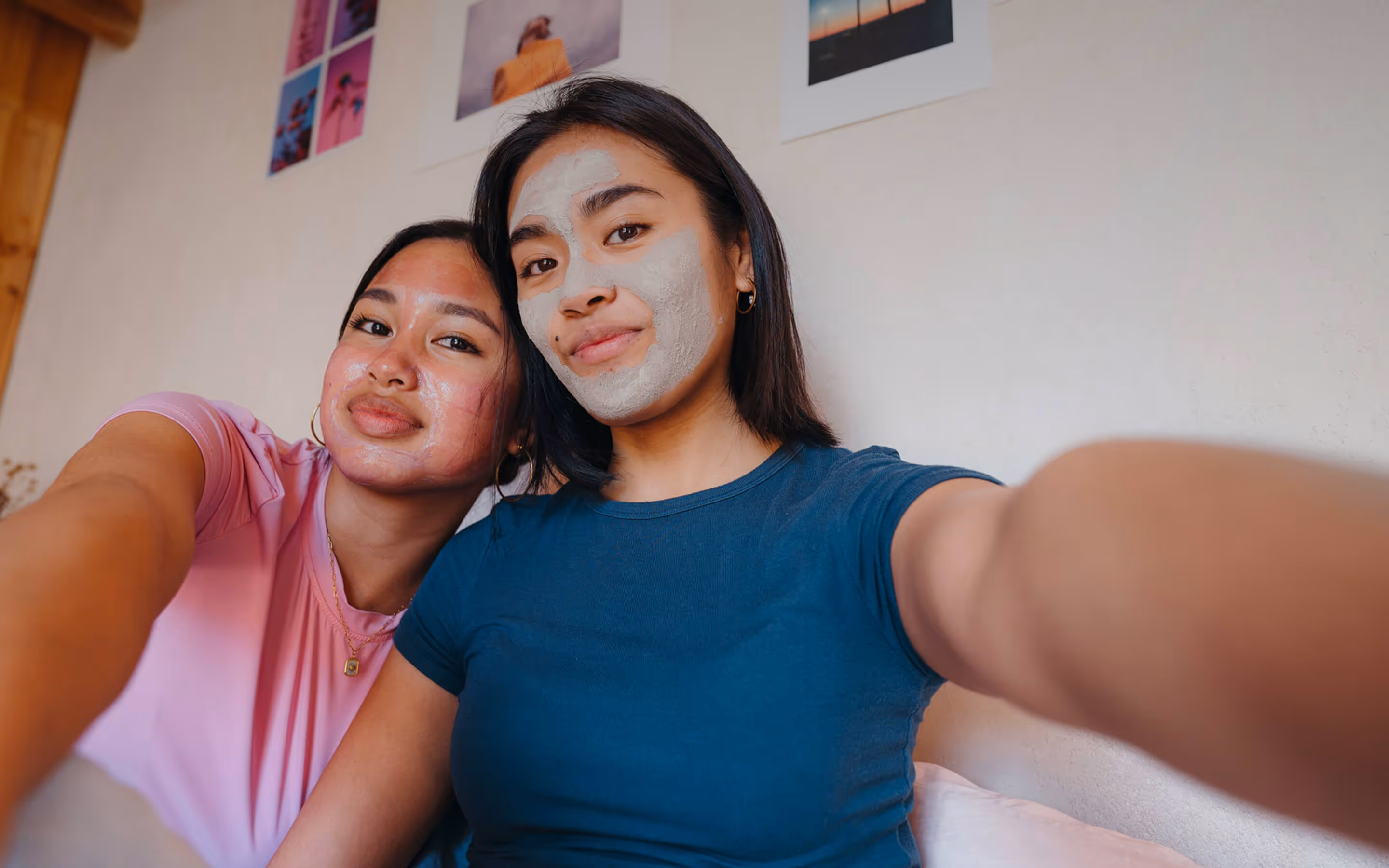 Dos mujeres jóvenes se toman una selfie de cerca juntas en el interior, ambas sonriendo suavemente mientras usan máscaras faciales, una con una máscara de gel rosado y la otra con una máscara de arcilla gris. Se sientan una al lado de la otra contra una pared de color crema decorada con coloridos estampados artísticos, lo que crea un ambiente acogedor y relajado que sugiere pasar un día de cuidado personal o de spa en casa.