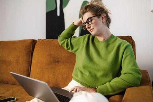 Una mujer con un suéter verde brillante y gafas se sienta en un sofá marrón, apoyando la cabeza en una mano mientras mira una computadora portátil. Su expresión relajada sugiere que está pensativamente comprometida con algo en la pantalla.