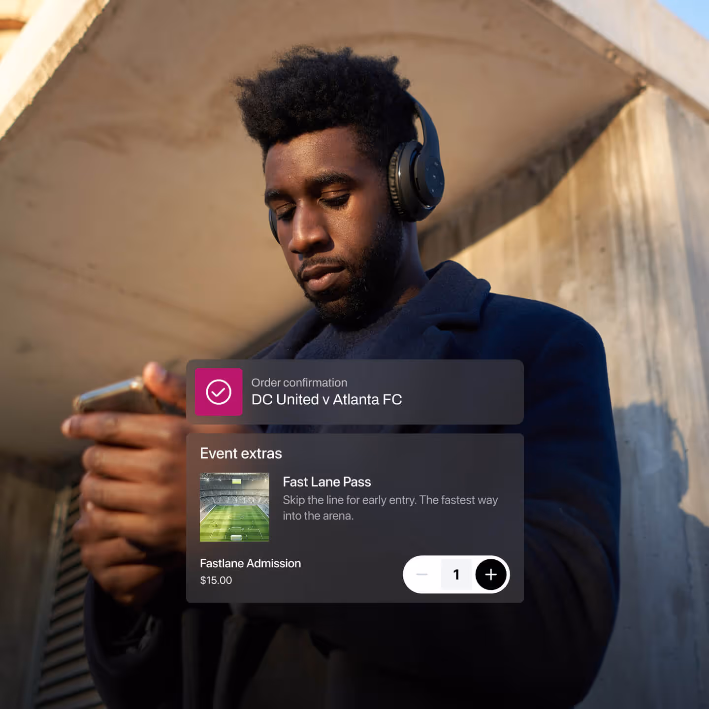 ワイヤレスヘッドフォンとダークコートを着た男性が、コンクリートの建築物の下に屋外に立ち、スマートフォンに焦点を合わせています。サッカーの試合の注文確認オーバーレイ「DC United v Atlanta FC」と、列をスキップできる「ファストレーンパス」のアップセルが表示されています。価格は15ドルで、利便性とイベント特典に重点を置いています。