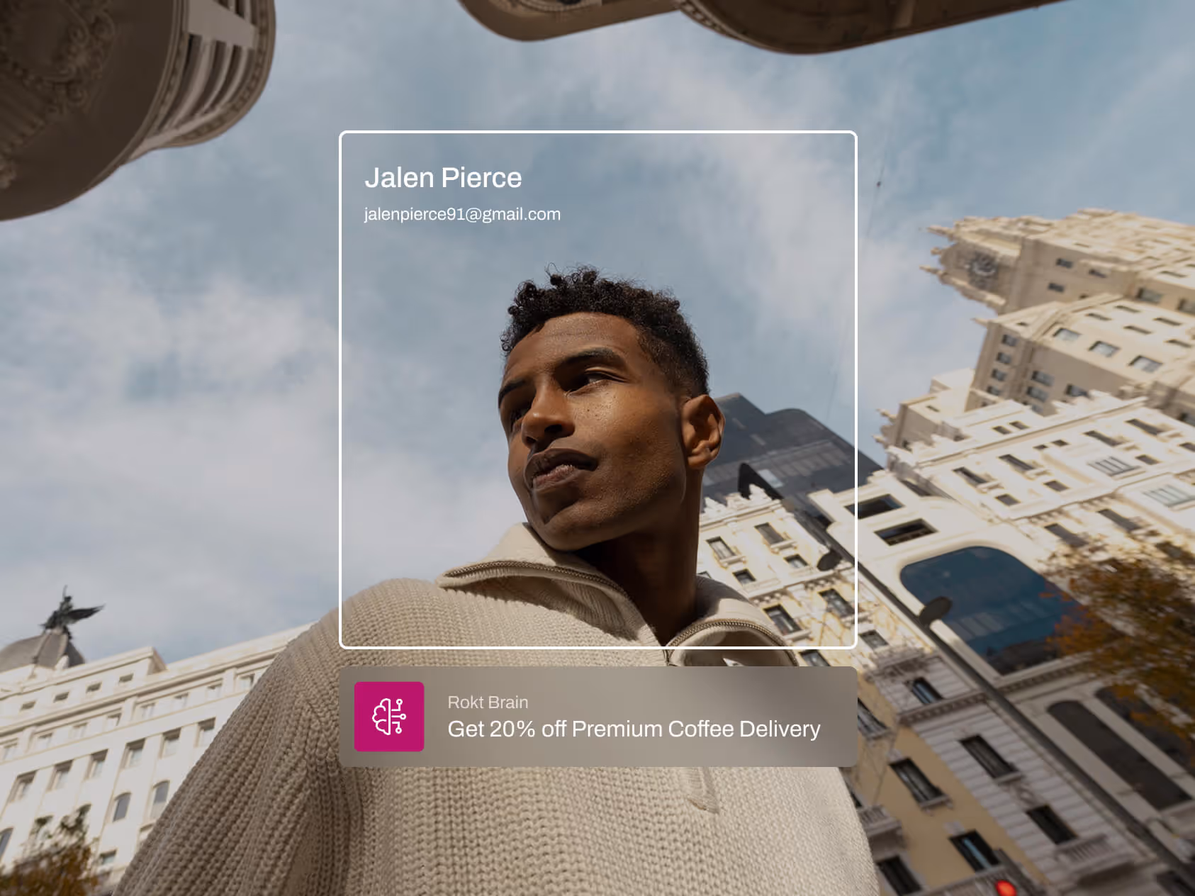 A man in a beige knit sweater stands in a city plaza, looking off thoughtfully beneath ornate architecture and a partly cloudy sky. A digital overlay with his name and email appears beside a promotional message offering 20% off premium coffee delivery from “Rokt Brain,” highlighting personalized email-based targeting.