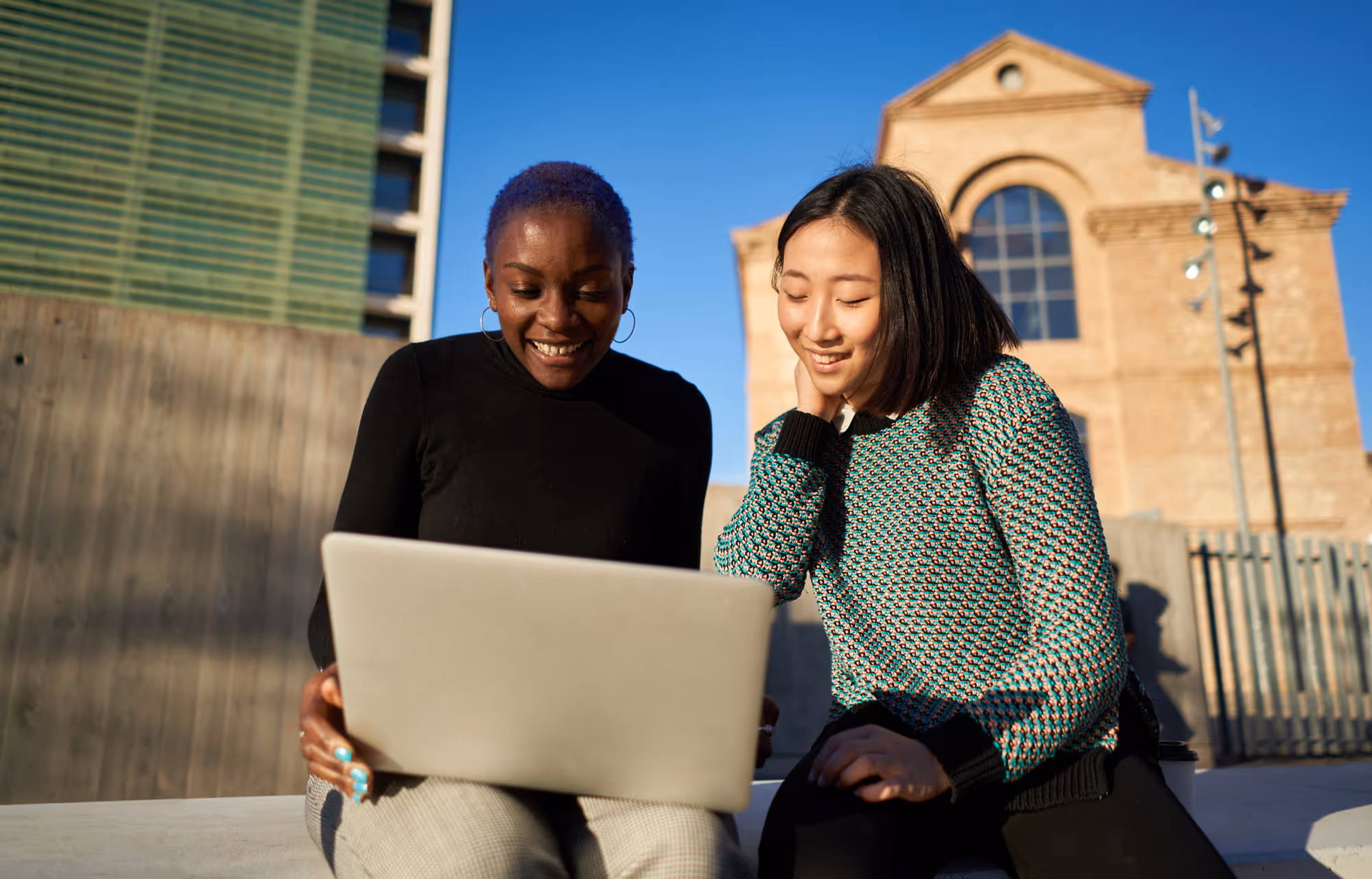 Two young women sitting outdoors looking at a laptop and smiling with a building and clear blue sky in the background.