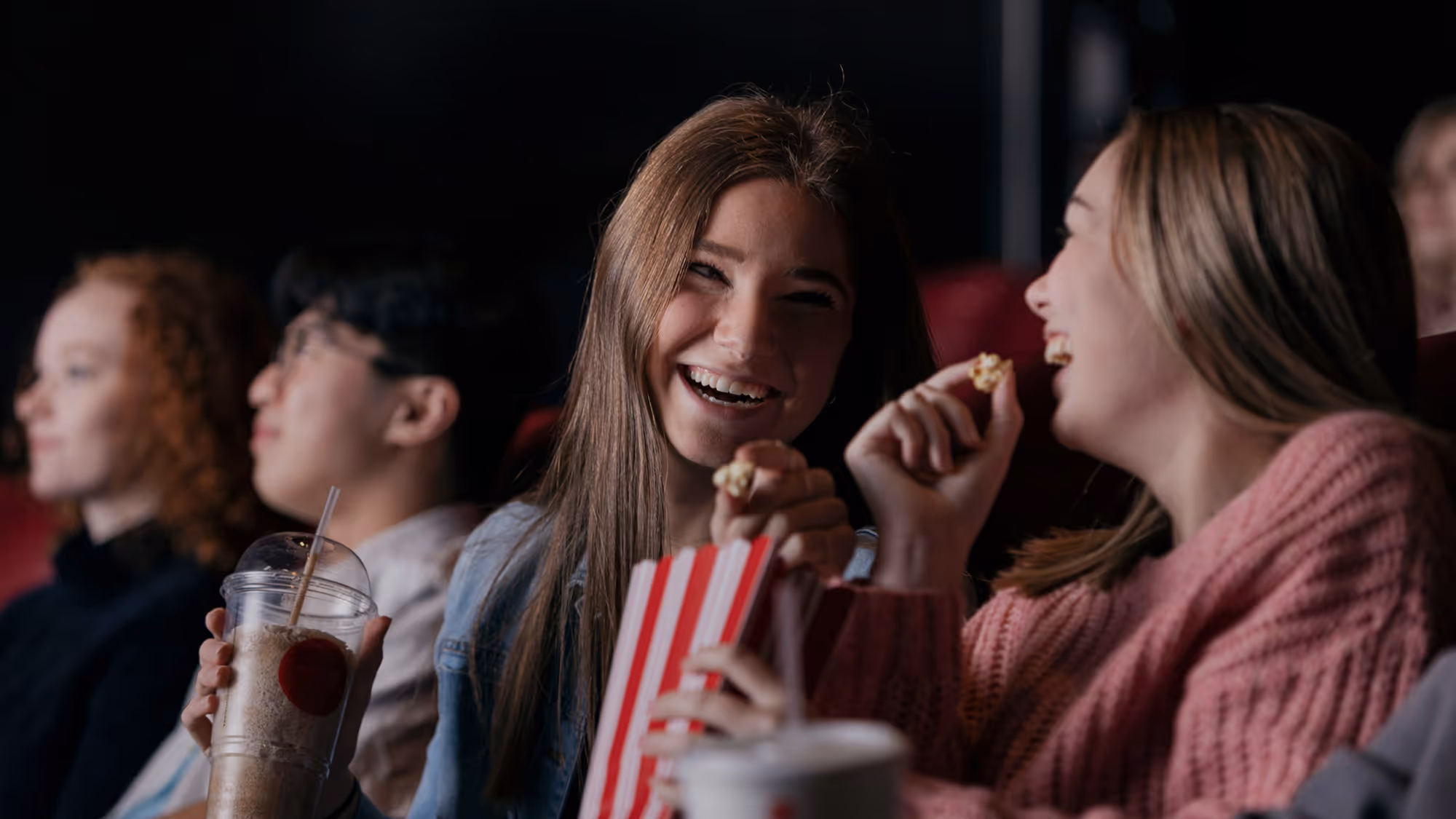 Two young women laugh and share popcorn while watching a movie in a theater, surrounded by other viewers. One holds a striped popcorn bag and the other a soda, capturing a candid, joyful moment of friends enjoying a night out.
