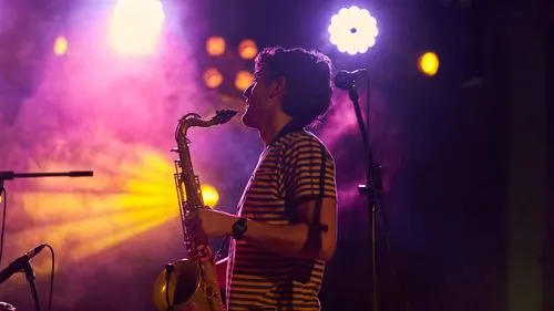 A saxophonist in a striped shirt plays on stage under dramatic purple and yellow lighting, with stage smoke and bright spotlights creating a moody, energetic atmosphere. A microphone stand is positioned beside him.