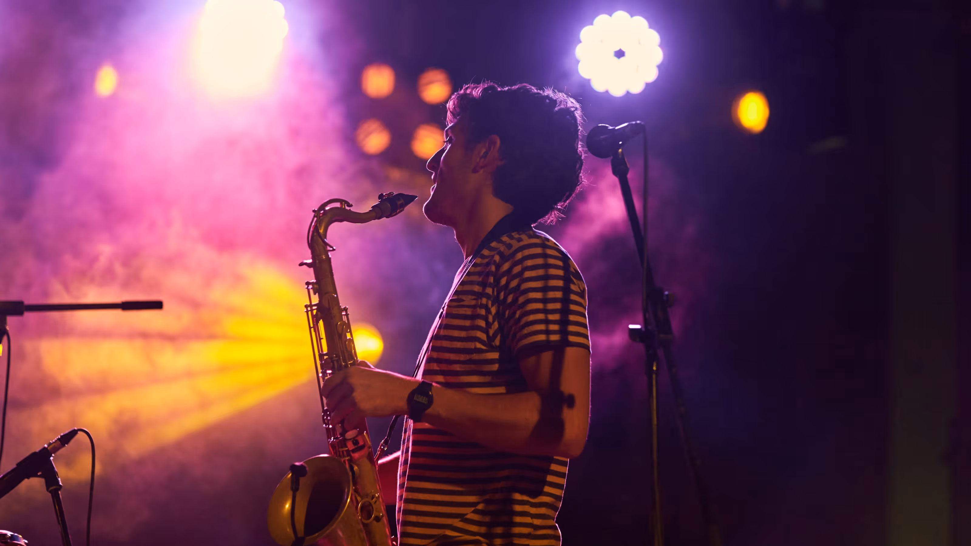 A saxophonist in a striped shirt plays on stage under dramatic purple and yellow lighting, with stage smoke and bright spotlights creating a moody, energetic atmosphere. A microphone stand is positioned beside him.