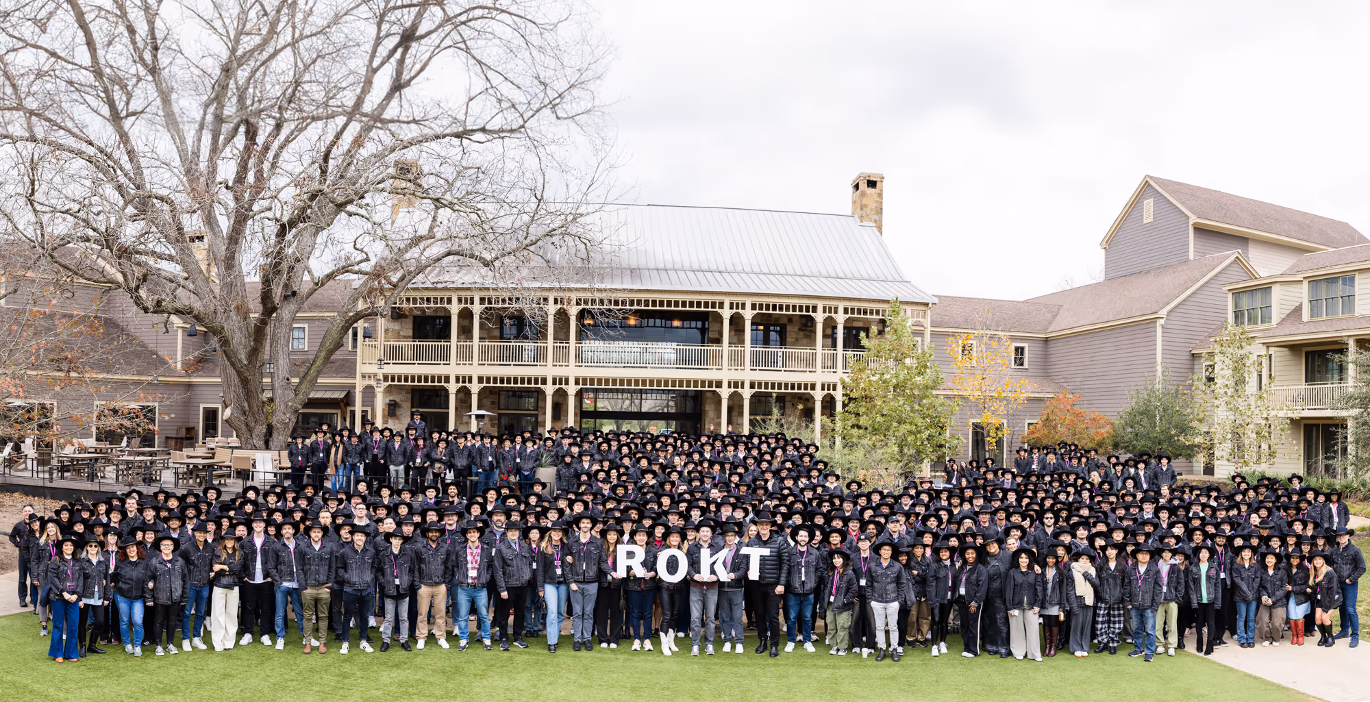 Grande photo de groupe de centaines de personnes debout en rangées sur une pelouse devant un bâtiment rustique de style lodge avec un revêtement gris et un toit métallique. Presque tout le monde porte un chapeau de cowboy noir et des vestes en jean foncées, créant un look cohérent et thématique. Au premier rang, plusieurs personnes brandissent des lettres blanches orthographiées « ROKT », indiquant qu'il s'agit d'un événement d'entreprise ou d'une réunion d'équipe. L'atmosphère est décontractée et joyeuse, certains souriant et posant en toute confiance. Derrière le groupe, des arbres sans feuilles et des feuillages automnaux épars suggèrent que la photo a été prise à la fin de l'automne ou au début de l'hiver.