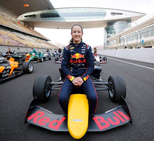 A female race car driver in a Red Bull racing suit sits confidently on the nose of a black and yellow Formula 4 race car with "Red Bull" branding on the front wing. She is smiling and looking directly at the camera. Behind her, other open-wheel race cars and drivers line up on the grid at a professional racetrack, likely before a race or media event. The modern grandstand and architecture in the background suggest a major international racing venue, possibly in Asia.