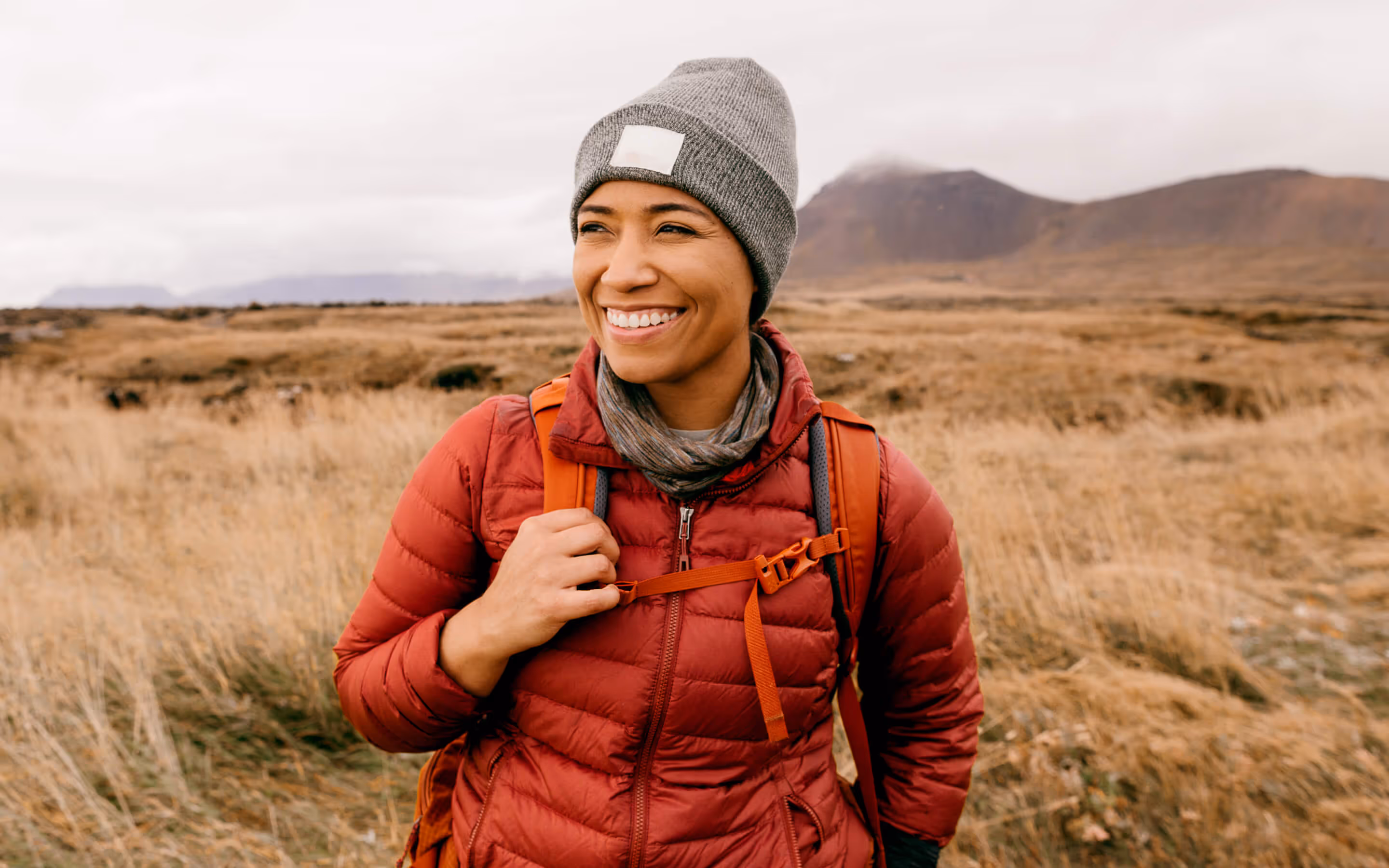 Smiling woman wearing a gray beanie and red jacket with an orange backpack in an open grassy field with mountains in the background.
