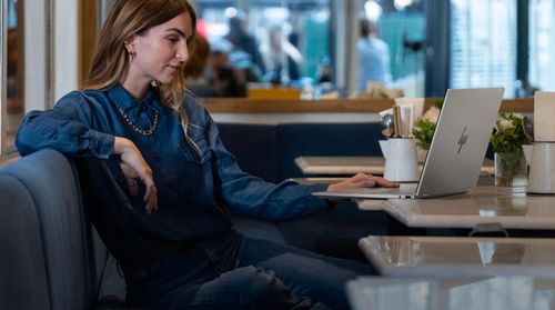 Woman in a denim shirt sitting at a cafe booth working on a silver HP laptop.
