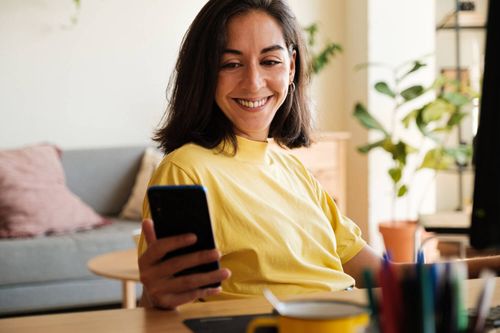 Una mujer sonriente con una camisa amarilla mira su teléfono inteligente mientras está sentada en una sala de estar acogedora y bien iluminada con plantas y muebles suaves en el fondo.