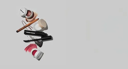 クリームブラッシュ、マスカラ、アイライナーペンシル、リップティント、バームなど、ジョーンズロードのメイクアップ製品をライトグレーの背景に平らに敷き詰め、各アイテムの横に色見本が巧みに塗られています。