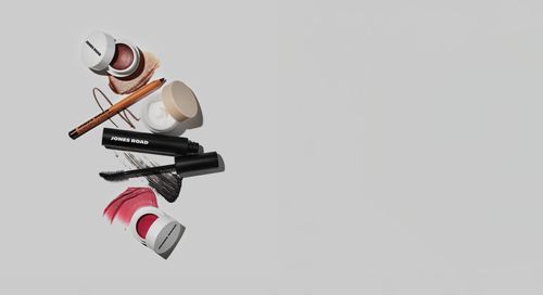 クリームブラッシュ、マスカラ、アイライナーペンシル、リップティント、バームなど、ジョーンズロードのメイクアップ製品をライトグレーの背景に平らに敷き詰め、各アイテムの横に色見本が巧みに塗られています。