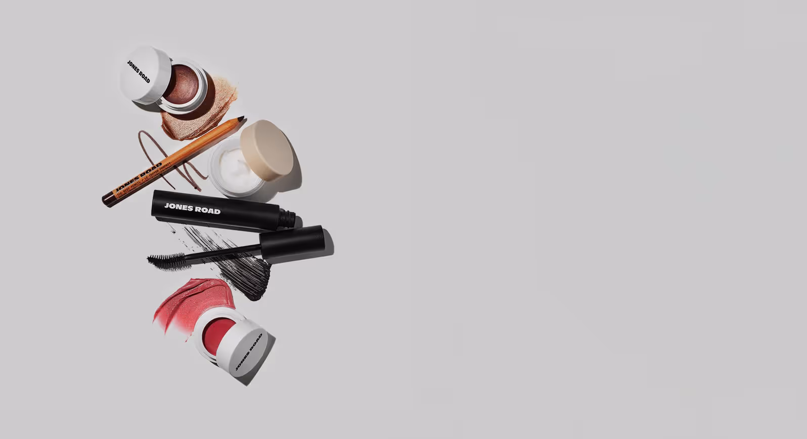 A flat lay of Jones Road makeup products on a light grey background, including cream blush, mascara, eyeliner pencil, lip tint, and balm, with color swatches artfully smeared beside each item.