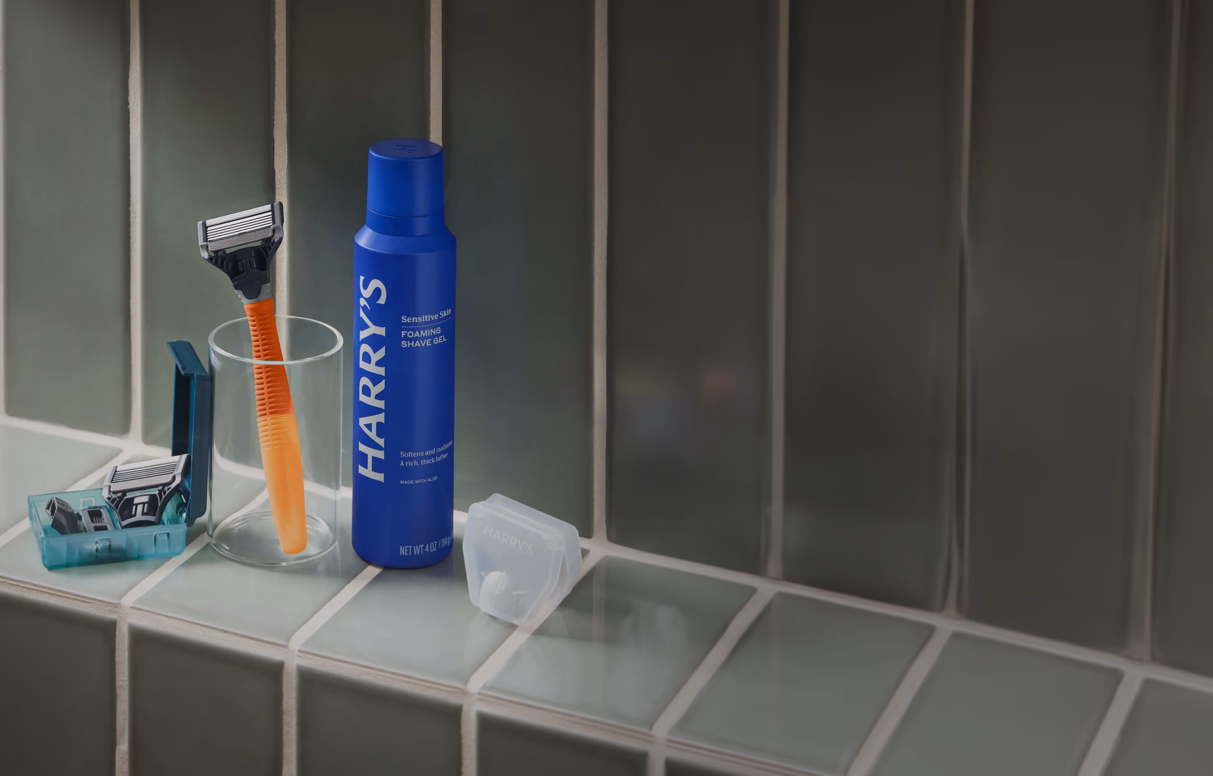A set of men's shaving products on a tiled bathroom shelf, including an orange-handled razor, blue shaving gel can labeled "HARRY'S," extra razor cartridges, and a plastic blade cover.