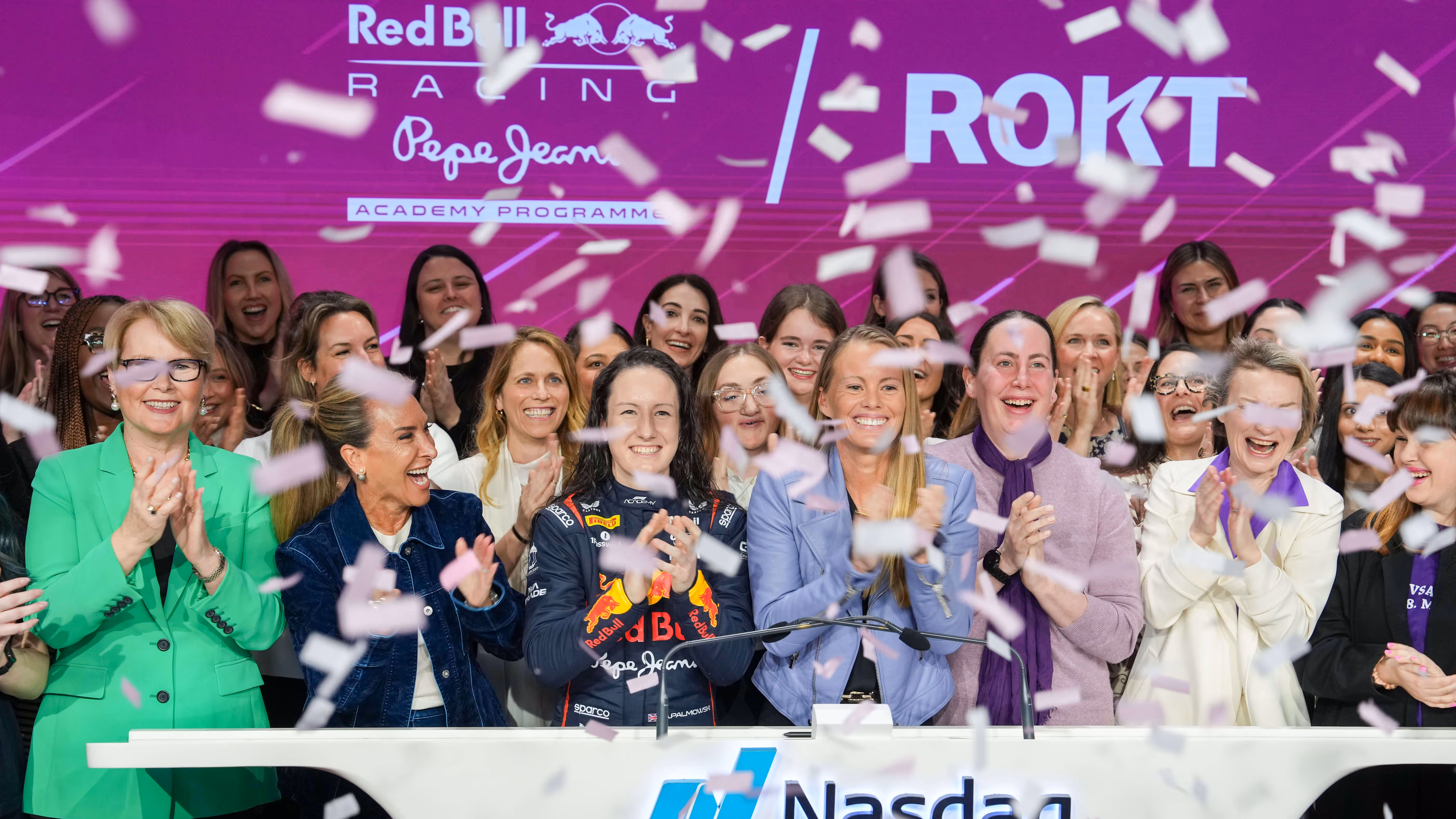 Un grand groupe de femmes fait la fête au milieu des confettis qui tombent sur le podium du Nasdaq, avec un pilote Red Bull Racing en uniforme au centre. La toile de fond présente les logos de Red Bull, de ROKT et du « Pepe Jeans Academy Programme », soulignant un événement marquant.