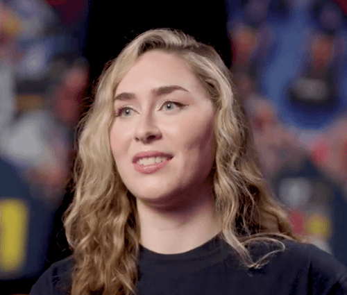 A woman with long, wavy blonde hair and a confident smile looks slightly off-camera against a blurred, colorful background. She wears a dark top and appears to be mid-interview or in a media feature setting.