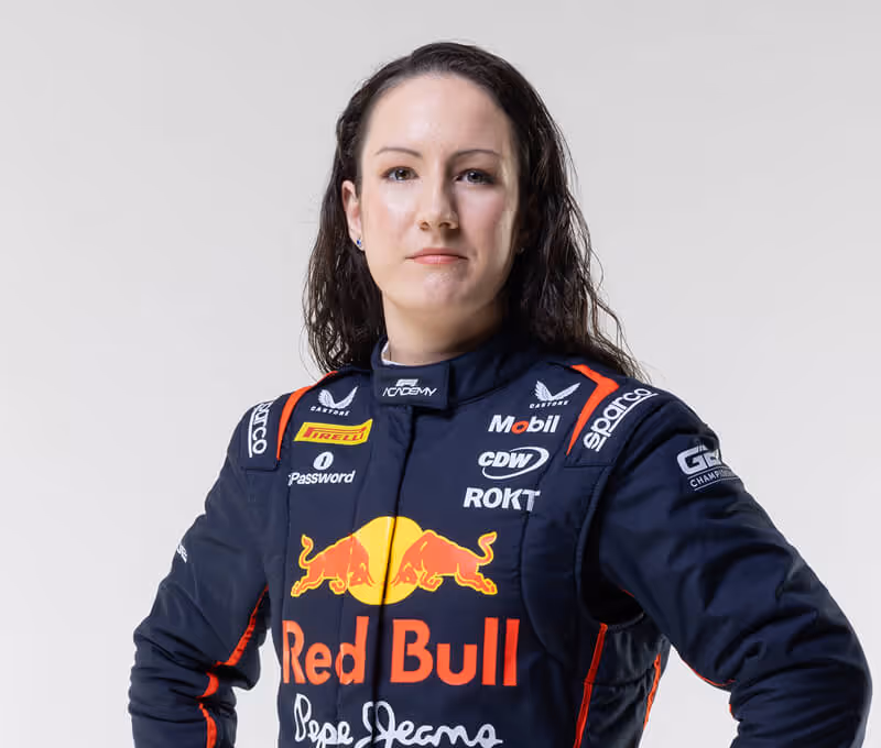 Pilote de course féminine dans une combinaison Red Bull Racing bleu marine, debout avec confiance sur un fond gris clair. Son costume arbore des logos de sponsors tels que ROKT, Mobil et Sparco, avec le logo Red Bull bien en évidence au centre.