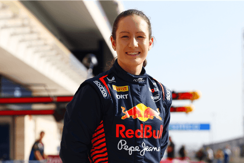 Smiling Red Bull racing driver standing in a sunlit pit lane, wearing a dark blue race suit with sponsor logos like Pepe Jeans, Pirelli, and ROKT. The blurred background includes garage structures and a few team members.