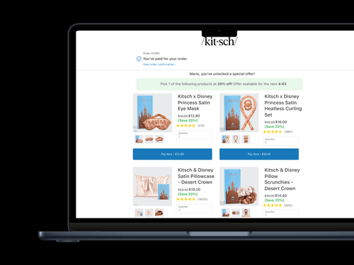ノートパソコンの画面には、ディズニープリンセスをテーマにした製品ラインを特集したキッチュの購入後のアップセルページが表示されます。上部には、注文 #1369 の支払いを確認するメッセージが表示され、その後に「Maria、スペシャルオファーがアンロックされました!」と書かれたパーソナライズされたバナーが表示されます。20% の割引カウントダウン付き。サテンアイマスク、ヒートレスカーリングセット、サテンピローケース、ピローシュシュの4つの商品が展示されています。それぞれ温かみのある砂漠の色調で、「デザートクラウン」というラベルが付いています。価格にはディスカウント、星評価、各アイテムの下に「今すぐ支払う」ボタンが表示されます。