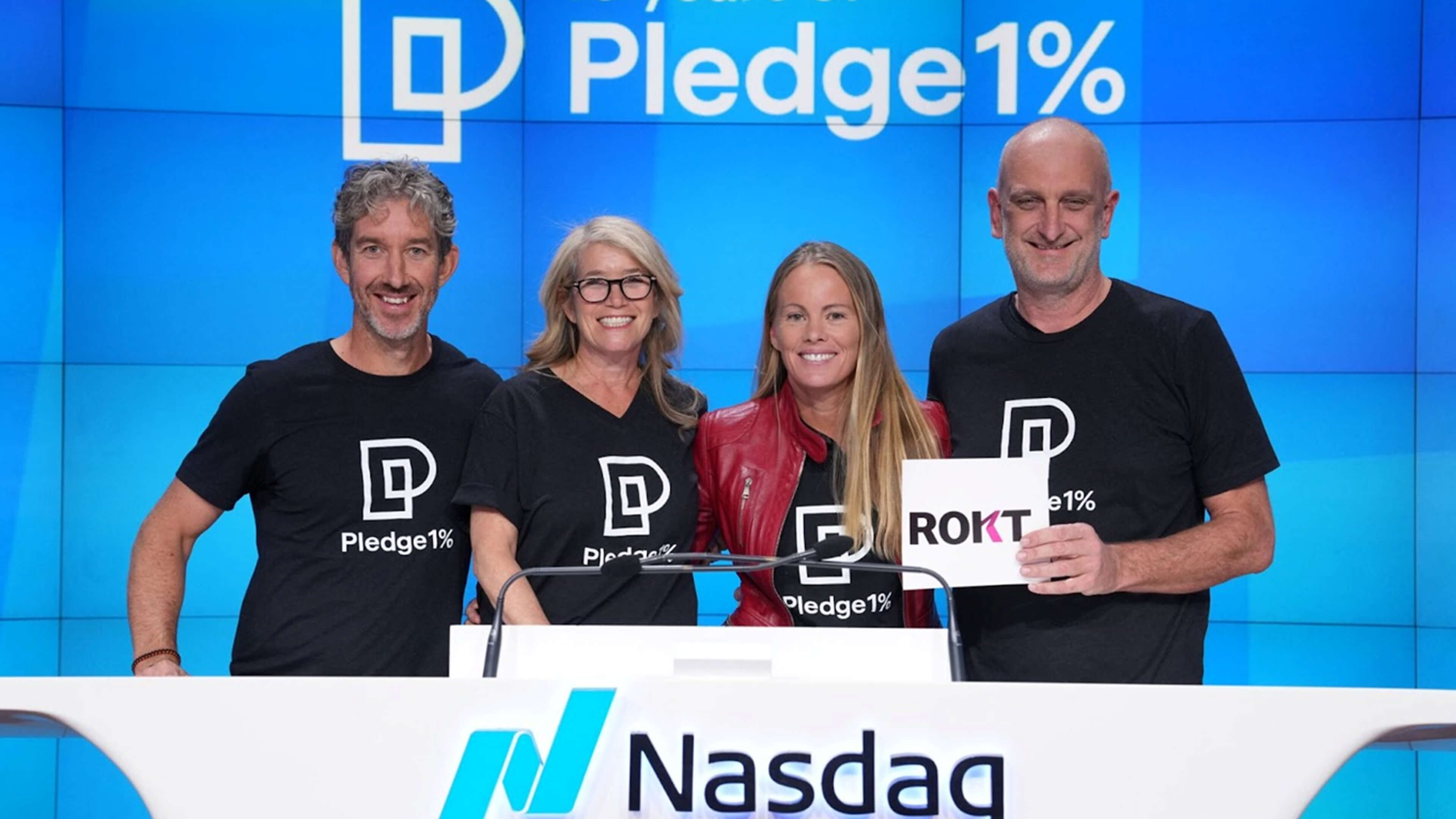 Four people stand smiling behind the Nasdaq podium during a celebratory event for Pledge 1%, a corporate philanthropy movement. All four wear black Pledge 1% t-shirts, except the woman second from the right, who pairs hers with a red leather jacket. The man on the far right holds a sign that reads “ROKT” in bold letters. Behind them, a large digital screen displays the Pledge 1% logo and name in white against a vibrant blue background, reinforcing the event’s branding and tech-forward setting.