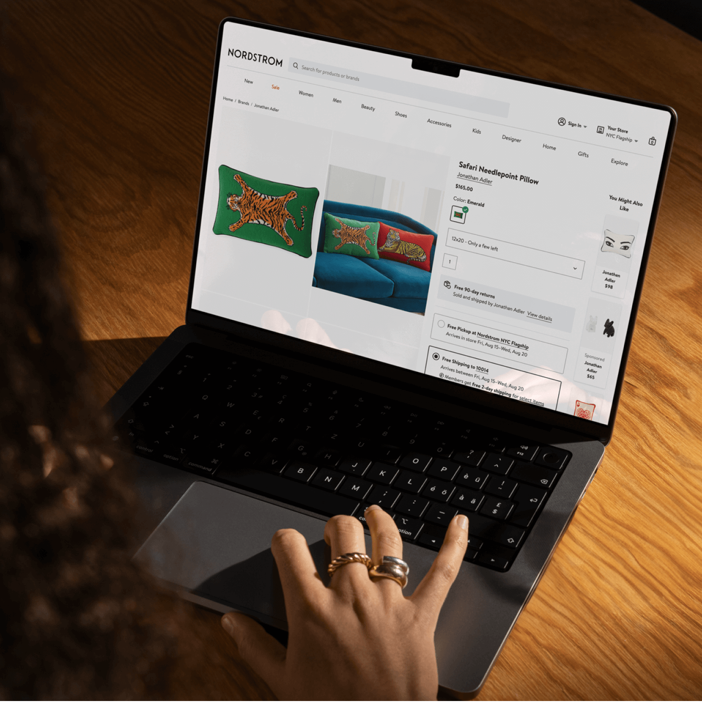 A person browses the Nordstrom website on a laptop, viewing a product page for a Safari Needlepoint Pillow by Jonathan Adler. The pillow features a bold embroidered tiger on emerald green fabric, shown styled on a bright blue couch. The hand on the keyboard, adorned with chunky gold rings, suggests a casual but stylish shopper engaging with an intuitive and visually rich ecommerce interface.