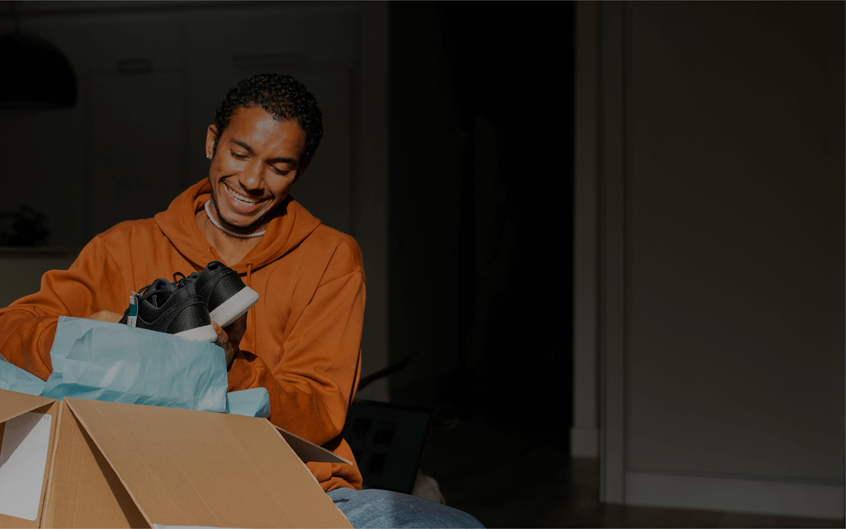 A young man wearing an orange hoodie smiles brightly as he unboxes a new pair of black sneakers with white soles. Sitting in a warmly lit room, he pulls the shoes out of a cardboard box lined with light blue tissue paper, clearly excited by the delivery. The background is dim, keeping the focus on his joyful reaction and the product.