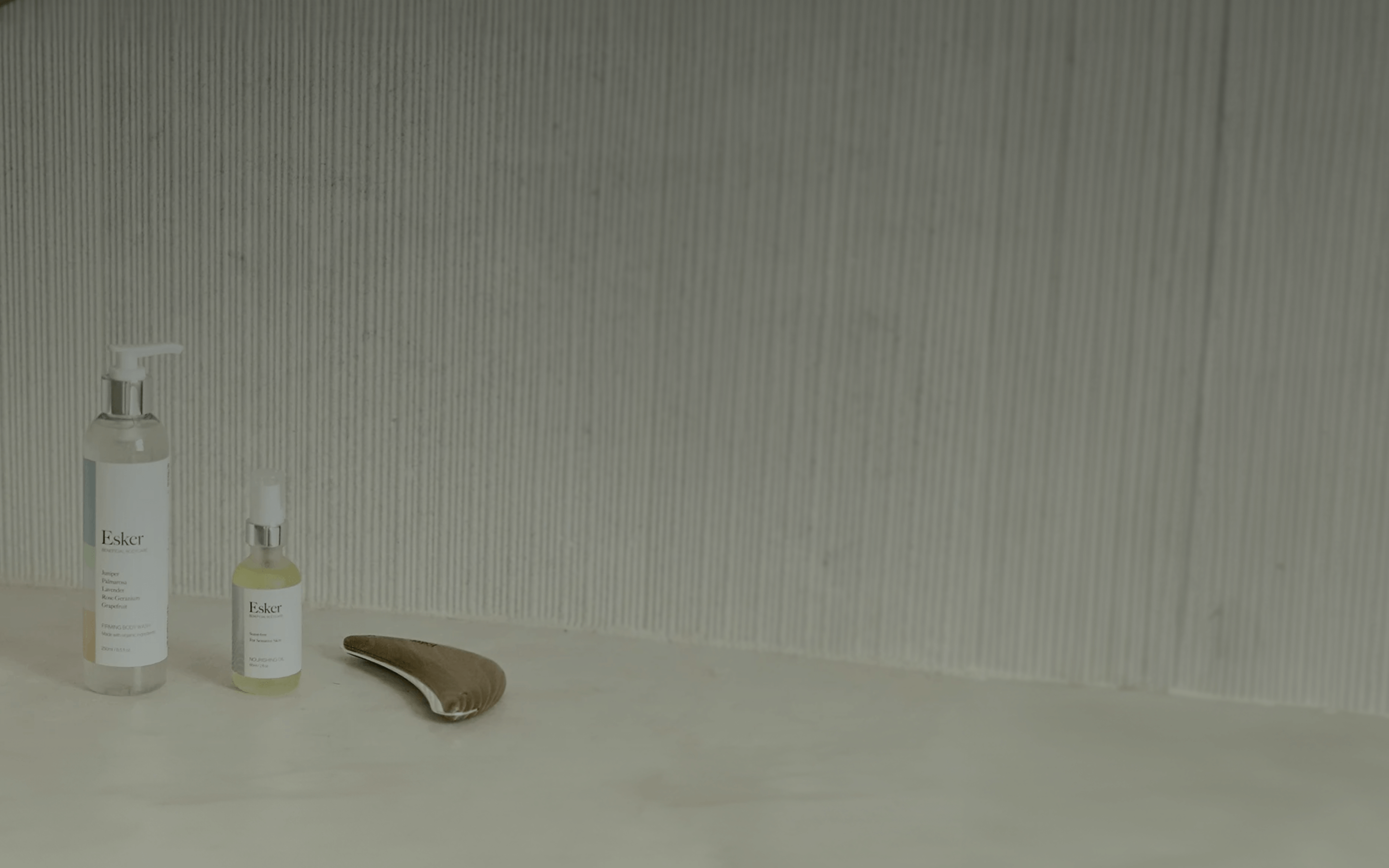 A minimalist skincare setup featuring two Esker brand products—a tall clear pump bottle and a smaller dropper bottle—arranged neatly on a light beige countertop. Beside them lies a wooden gua sha tool with a smooth, curved shape. The backdrop is a vertically ridged gray wall, creating a clean, spa-like atmosphere that emphasizes calm and self-care.