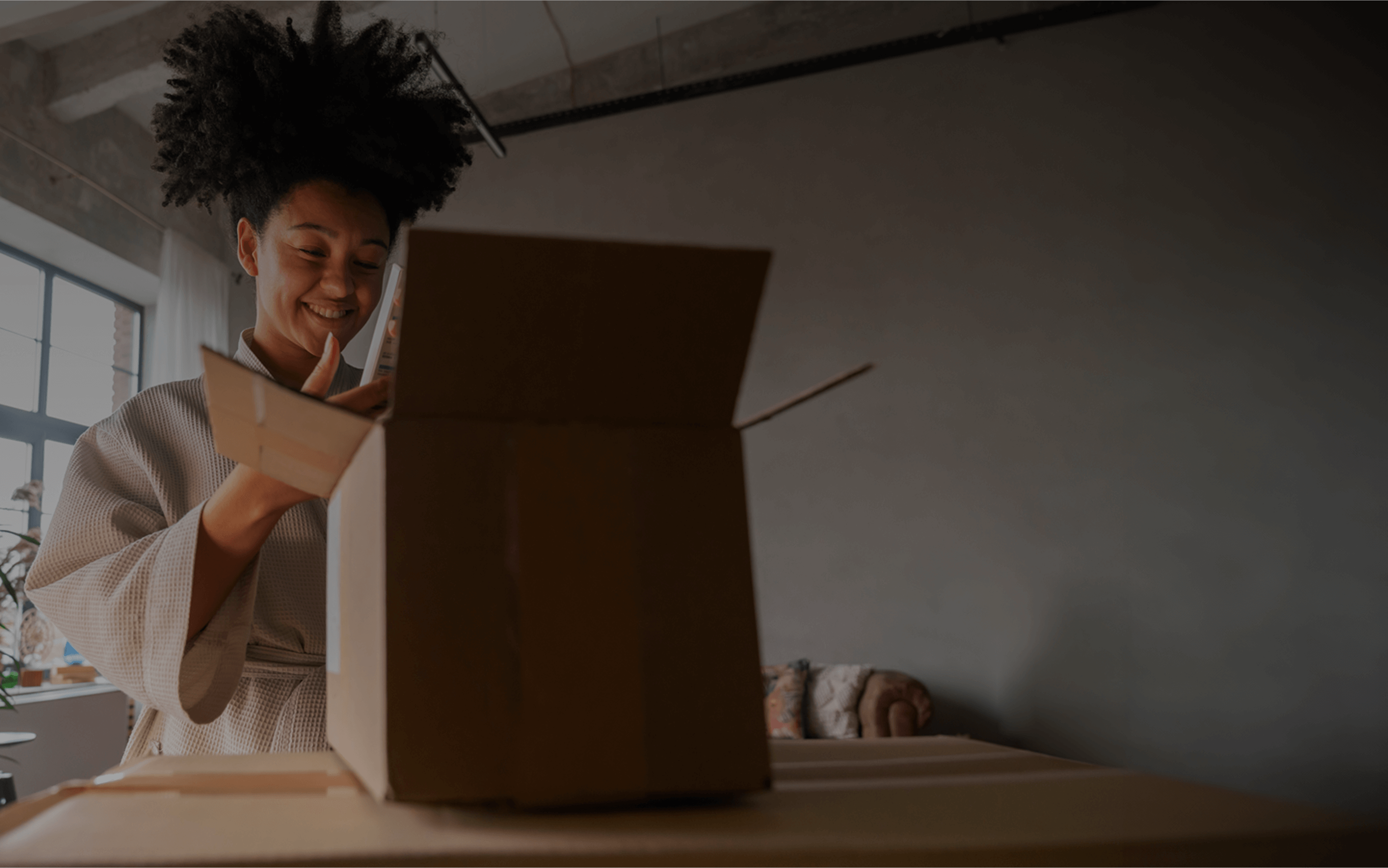 A woman with a joyful expression stands in a cozy, softly lit room, opening a cardboard box placed on a table. Dressed in a textured bathrobe, she examines the contents inside with curiosity and a smile. Large windows let in natural light, brightening the industrial-style space with exposed beams and a casual, lived-in feel.