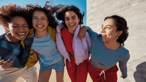 Four diverse young women laughing and embracing outdoors against a blue sky and concrete wall.
