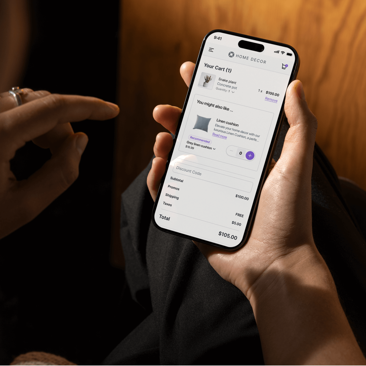 A person holds a smartphone displaying an online shopping cart for a home decor app. The cart contains a “Snake plant in a concrete pot” priced at $100, with a recommendation below for a “Linen cushion” in grey for $18.99. The subtotal is $100, with free shipping, $5 in taxes, and a total of $105. A second hand is poised to interact with the screen, possibly to add the cushion. The setting appears cozy and dimly lit, suggesting relaxed evening shopping.