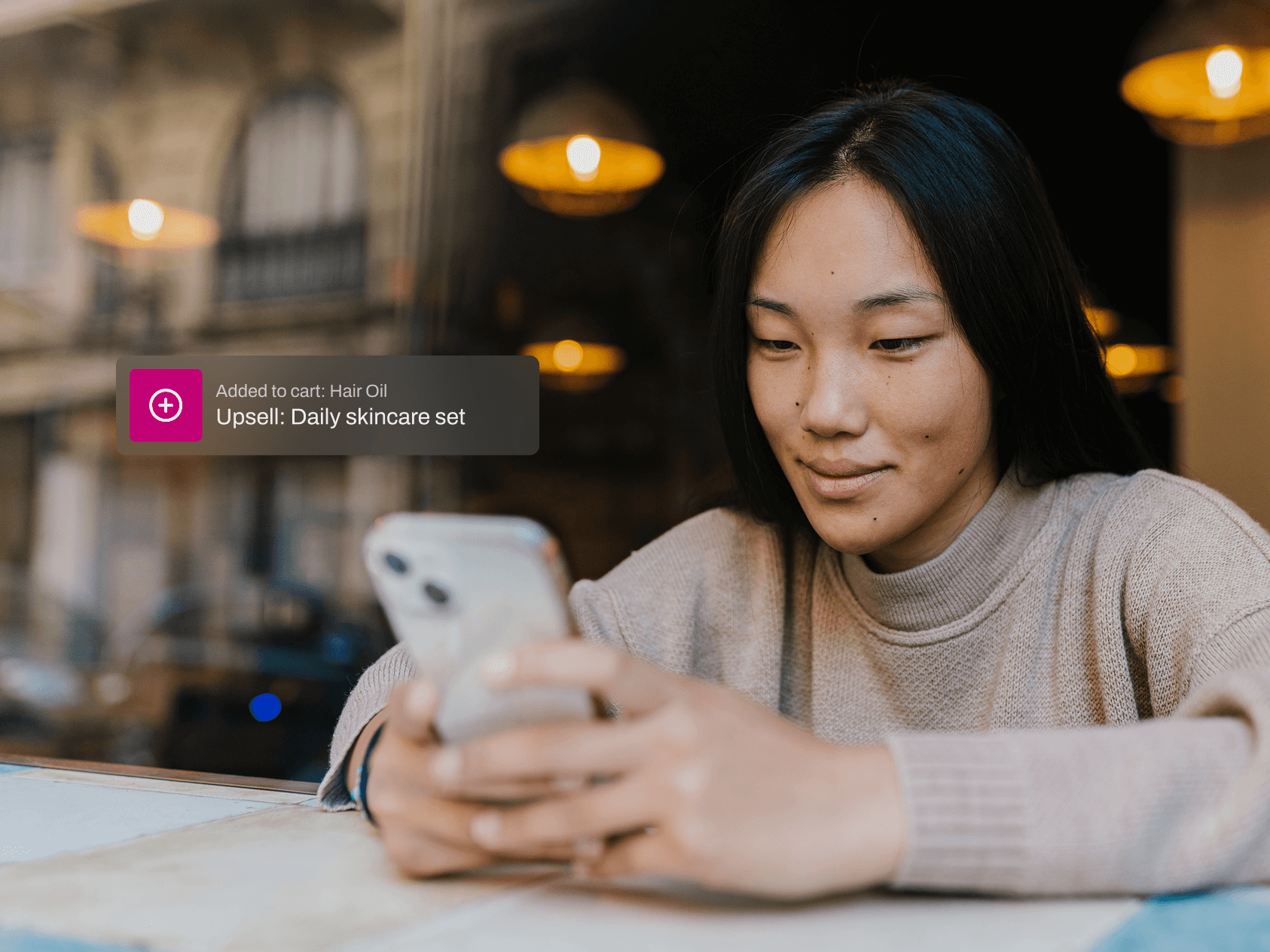 A young woman sits by a window in a cozy café, smiling slightly as she looks at her smartphone. She wears a beige knit sweater and has long black hair. On the screen, a shopping notification appears, reading: “Added to cart: Hair Oil” with an upsell prompt below it saying “Upsell: Daily skincare set.” Warm pendant lights and a blurred street view reflect through the glass behind her, creating a relaxed, modern shopping moment.