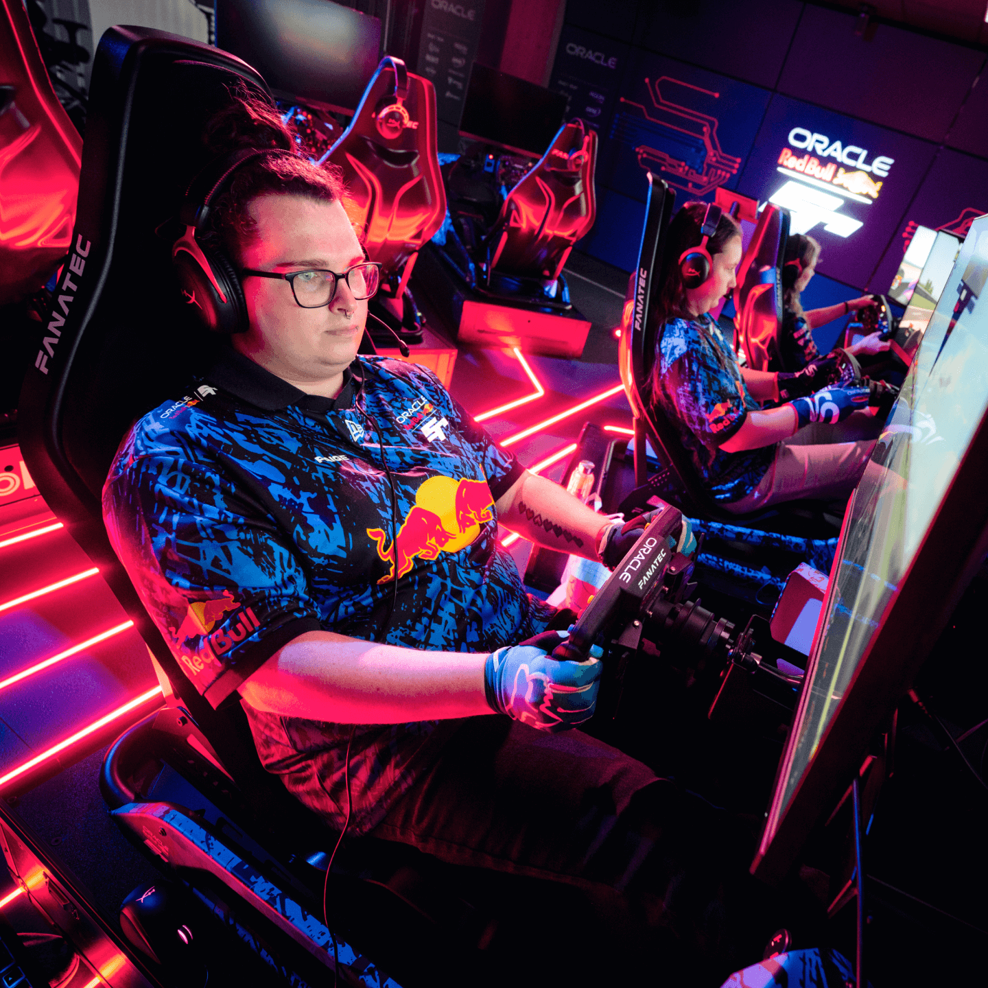 Three esports drivers wearing Red Bull Racing and FaZe Clan gear intensely compete in a racing simulator setup with steering wheels and monitors, surrounded by neon pink lighting in a high-tech gaming arena.