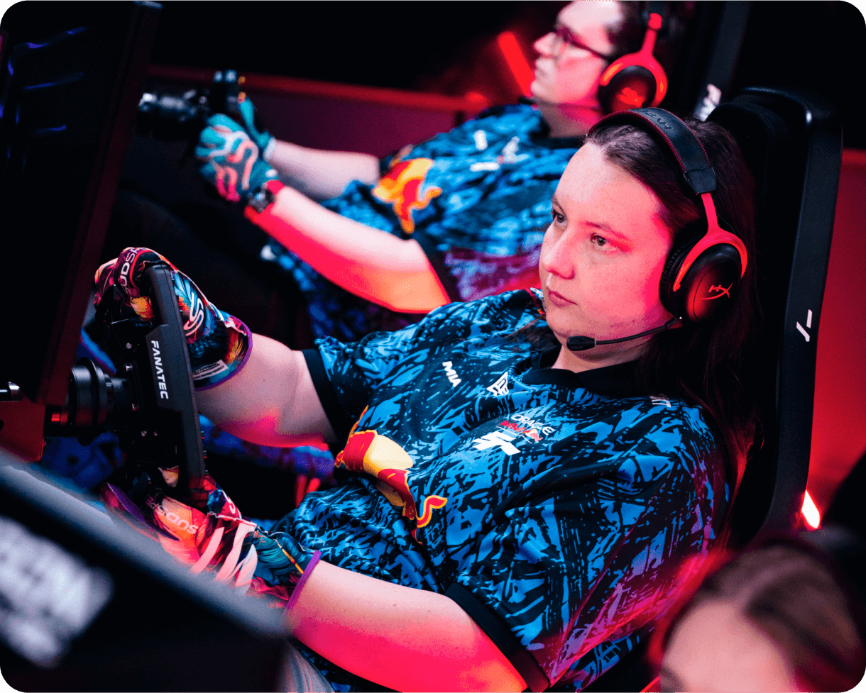 A focused esports racer grips a Fanatec racing wheel in a simulator setup, wearing a Red Bull Racing jersey and gaming headset. Red and blue lighting highlights the intense atmosphere, with another player visible in the background.
