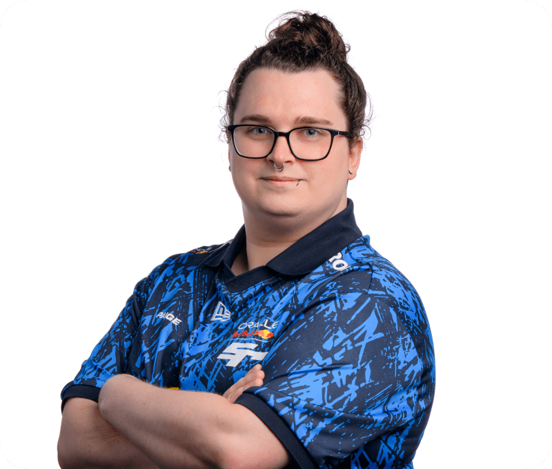 A confident individual with dark hair in a top bun, wearing glasses and a septum piercing, poses with arms crossed in a blue Oracle Red Bull Racing esports jersey. The background is plain white.
