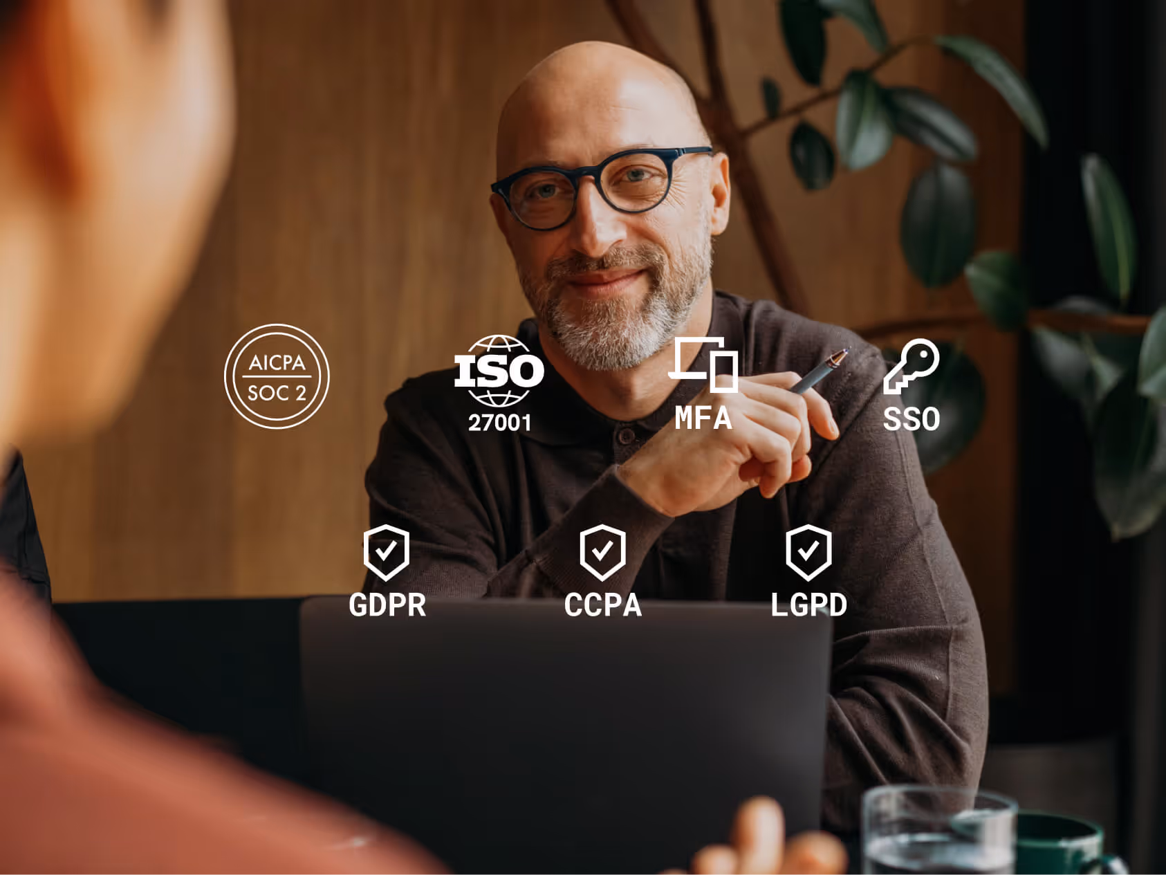 Smiling bald man with glasses holding a pen behind a laptop, digital security and compliance icons floating around him.