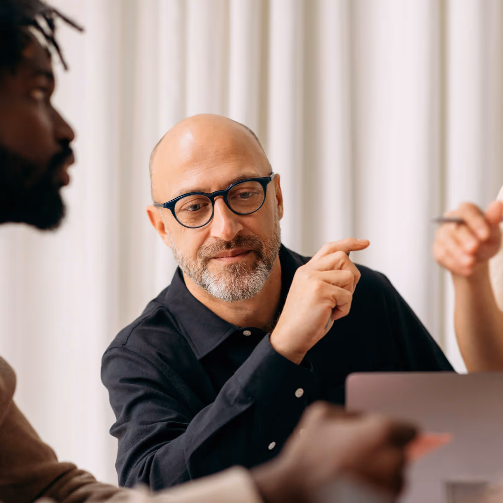 Bald man with glasses pointing at a laptop screen during a discussion with colleagues in a bright room.
