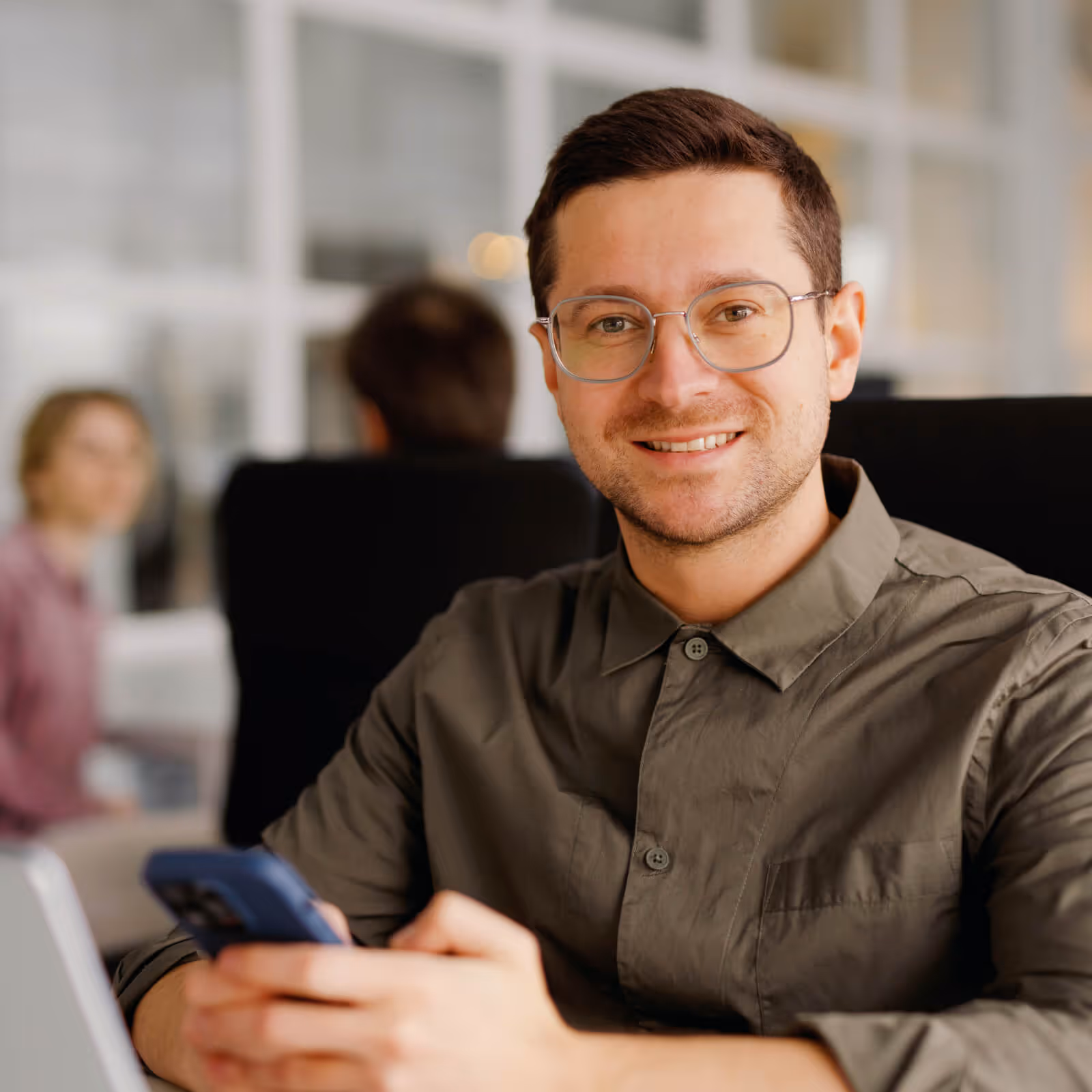 Smiling man with glasses holding a smartphone in an office setting with colleagues in the background.